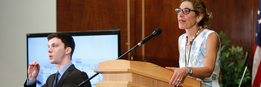 A woman speaks at a podium. A man stands beside her signing.