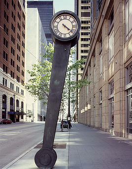 Color photo shows a tall black metronome-shaped clock on a city sidewalk. Clock is tilted slightly to the right.

