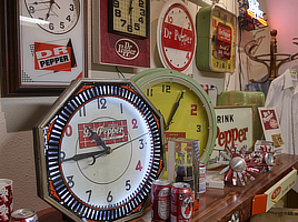 Close up photo of a wall filled with clocks of different shapes and colors, all advertising Dr. Pepper.
