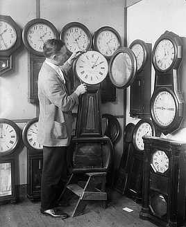 Black and white photo shows a thin white man standing in a room filled with more than 10 wall clocks with large dials.
