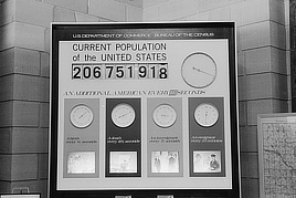 Black and white photo shows a large sign with several clock faces that represent  changes in population. The changes cover births, deaths, immigration, and emigration.
