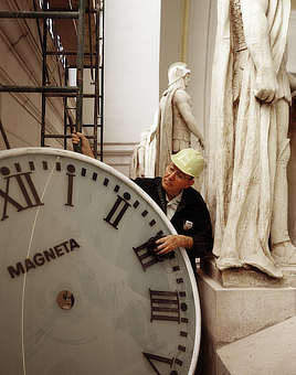 Color photo shows scaffolding and larger than life classical statuary. In the center, a man in a hard hat holds and cleans with a rag a clock face that is larger than himself.
