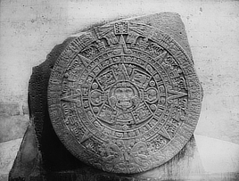 Black and white photo shows a large stone circle with intricately engraved concentric designs. In the center is a face of a deity.
