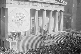 Photo shows a crowd in front of a large neoclassical building with a statue of George Washington. A large banner says: “Liberty Loan Clock, Make Your Dollars Fight For Freedom.”
