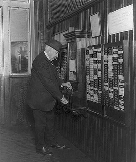A man in a dark hat and suit and a cigar in his mouth, stands in a dark paneled room. He maneuvers a small card into a machine hanging on the wall.
