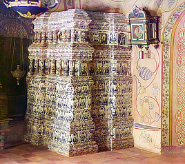 Room interior shows a wall clock and large ceramic tile heating stove with decorative frescoes on the walls.
