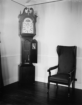 Tall clock and empty chair in a room corner. The clock case is open to show the pendulum and weights. Decorative finials at top.
