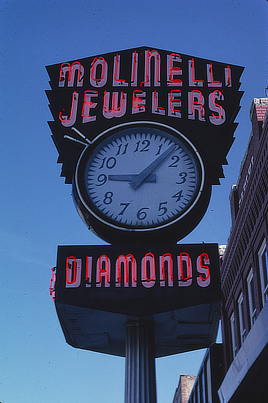 Photo shows a large street sign sitting on top of a tall pole. Neon red letters spell out the name of the business above a clock, and below it says “Diamonds.”
