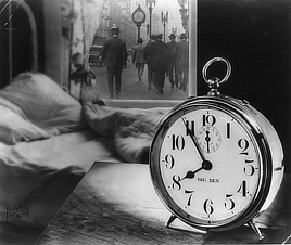 Black and white photo shows an alarm clock in the foreground, and an unmade bed behind it. A street scene with another clock appears through a window.
