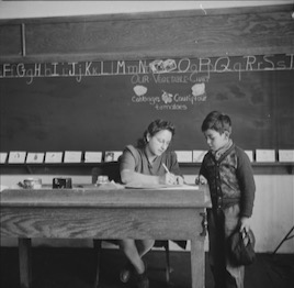 A young boy in a sweater stands at the edge of a desk where his teacher writes on paper. Behind them, a chalkboard displays the English alphabet and lists the names of various vegetables.