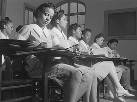 A group of women sits at student desks wearing matching uniforms.