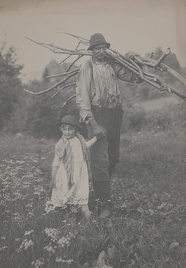 The man wears a ragged shirt and looks at the camera while holding the hand of a young child wearing a dress and shawl. Both wear caps.