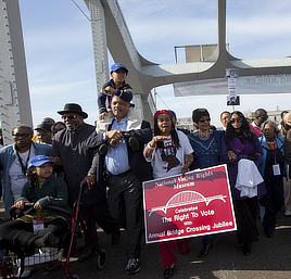 Crowd of Black men, women, and children stands on the bridge. One woman holds a sign that reads "National Voting Rights Museum. Celebrate the right to vote with annual bridge crossing jubilee."
