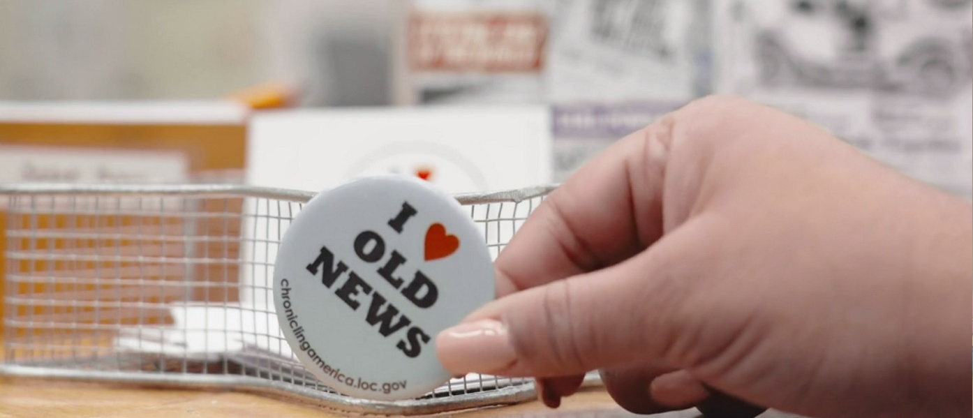 Photo of "I love old news" button being held between thumb and forefinger over a desk with metal basket in the background.
